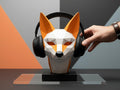 Happy3dWorld low-poly geometric fox headphone stand in orange and white PLA, holding black headphones on a desk.