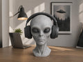 Happy3dWorld gray alien bust headphone stand holding black over-ear headphones on a wooden desk in a sci-fi room.